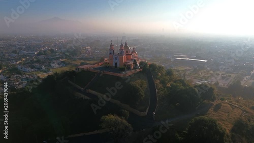Aerial drone orbits the hilltop church atop the Cholula pyramid at dawn, with the Puebla skyline spreading below and the Popocatepetl and Iztaccihuatl volcanoes rising on the horizon