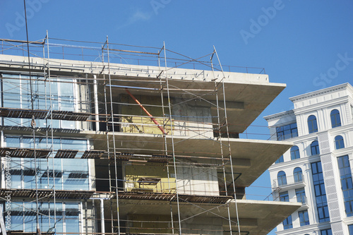 A close-up of a multi-story reinforced concrete residential or office building under construction. The facade is covered with steel scaffolding.
