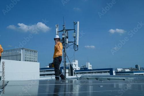 Technician pair in yellow safety jackets discuss work beside a rooftop telecom antenna on a rooftop solar farm, showing sustainable power, inspection and maintenance on a rooftop.