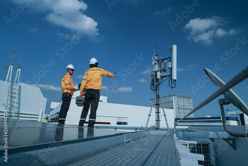 Quadro su tela On-site view as inspection pair in yellow jackets discuss work beside a rooftop telecom antenna overlooking the city, capturing renewable energy, green technology and engineering
