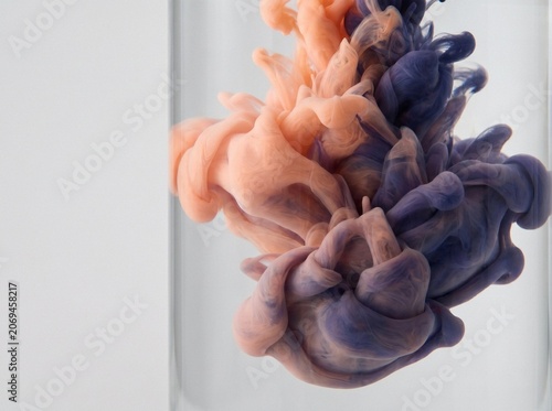 Abstract peach and purple ink swirling in clear water inside glass vessel against light background