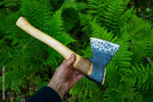 Epic handmade forged steel axe hatchet tool green fern forest background vertical view.
