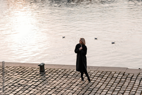 Photos Young woman in stylish dark coat standing on Vltava river embankment in Prague