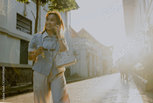Fashionable adult Asian woman take off his sunglasses talking by phone call walks on the alley city street at sunsets. Commuting, urban lifestyle and workwear fashion concept.