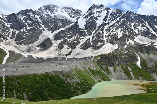 Fotografi Emerald mountain lake in the Caucasus Mountains during summer, surrounded by greenery with glacier-covered peaks and the Semerka glacier in the background