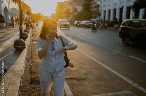 Fashionable style adult Asian Businesswoman in casual button down shirt talking by phone call looks at wristwatch while walking in city street at sunsets.