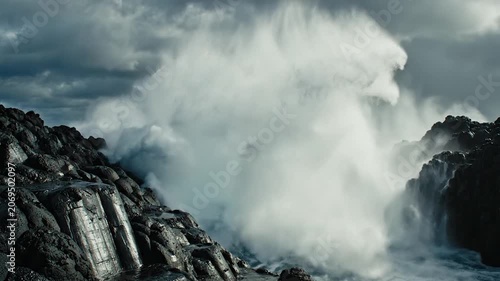 Powerful ocean waves crashing against dark volcanic rocks under a cloudy sky