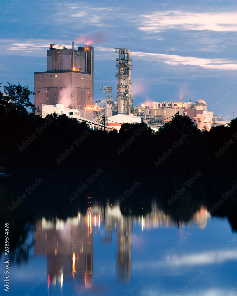 Naklejka premium Dramatic gritty and industrial paper mill reflecting on a quiet lake at blue hour