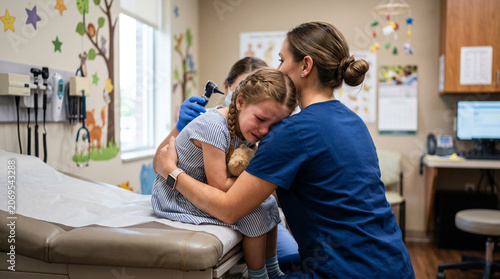 Pediatric nurse in dark blue scrubs hugging and comforting a crying young girl during a difficult ear examination by a doctor in a children's medical exam room, ideal for pediatric healthcare content.