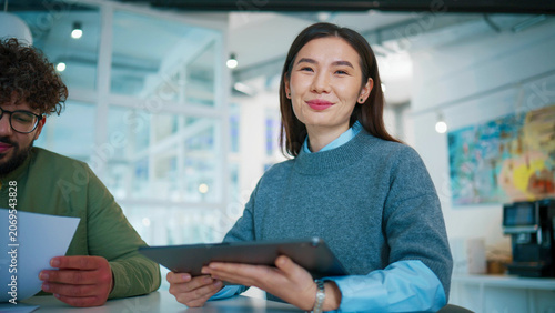 Professional woman holding digital tablet at desk. Confident female looking at camera. Male coworker checking printed report. Business colleagues working in bright glass office interior.
