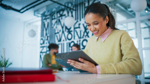 Creative office with abstract wall decor. Smiling female holding tablet and smiling gently. Reading content on screen. Standing near desk with documents. Woman reviewing project details during work.