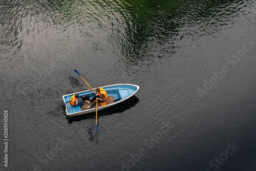 Overhead scene shows small blue rowing boat carrying two people in orange life vests moving across dark rippled water with oars extended