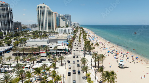 A cinematic aerial view captures the vibrant energy of Fort Lauderdale Beach, where turquoise Atlantic waters meet a lively beachfront lined with palm trees, hotels, and coastal activity.