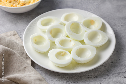 Hard boiled egg halves with yolks removed on a white plate, prepared for stuffed eggs recipe on a gray textured surface with soft natural light