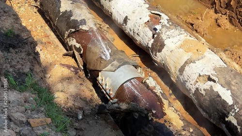 Two old heating pipes are exposed in an excavated trench while water leaks from one pipe. Concept: worn infrastructure, utility repair and emergency maintenance.