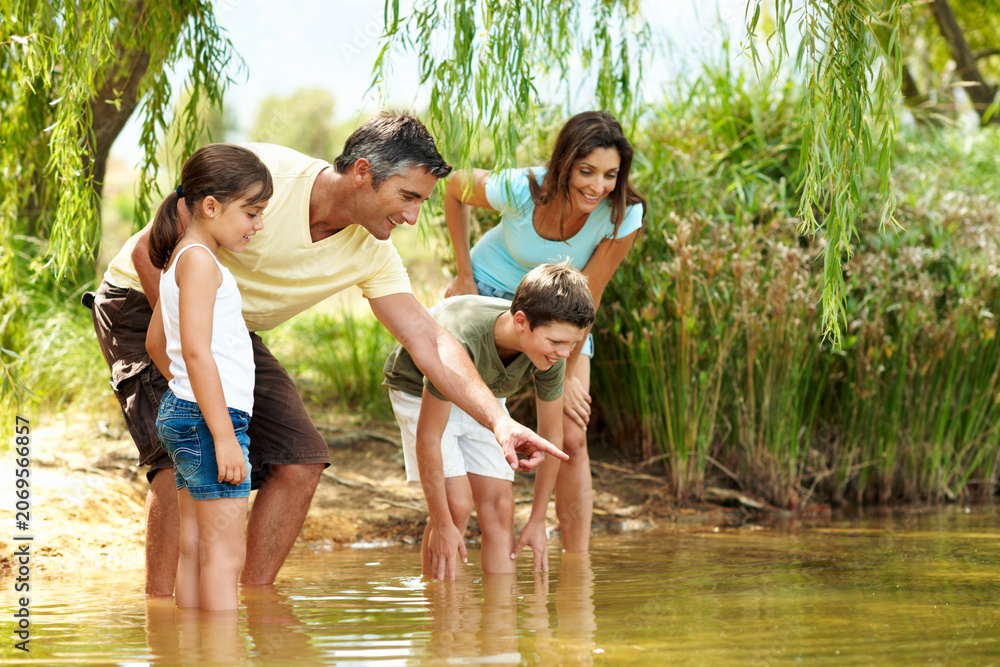 Obraz premium Happy, parents and pointing with children at lake, adventure and discover fish on holiday. Smile, people and hand gesture in nature with kids, river observation and water for sightseeing vacation