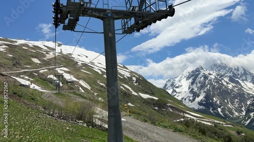 Mountain ski lift moves upward along snow-covered terrain, surrounded by green grass and towering peaks under a clear blue sky