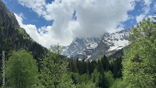 Lush green trees frame the majestic mountains under a cloudy sky, highlighting the serene beauty of a springtime landscape in nature