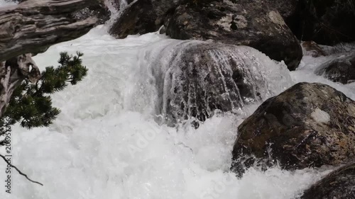 Water flows rapidly over smooth rocks in a stream, with splashes and bubbles visible, surrounded by lush greenery and natural textures
