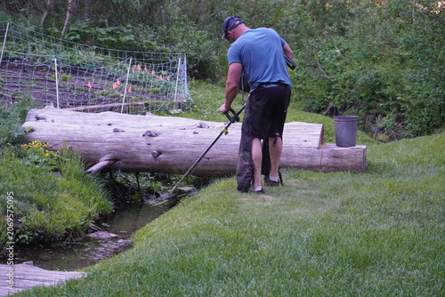 Back view of a man trimming overgrown grass and weeds with a string trimmer near a natural log bridge spanning a small stream in a lush backyard garden.