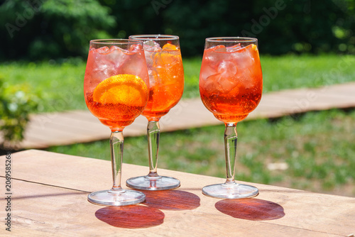 Three glasses of refreshing orange spritz cocktails with ice and citrus slices served outdoors on green sunny patio. Summer relaxation, outdoor dining, leisure, travel and social gathering concept.