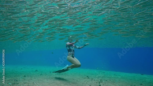 Young female freediver hovers beneath surface in eye of massive Silverside school. Fish bait ball form perfect halo that flows continuously around graceful girl snorkeler on shallow water
