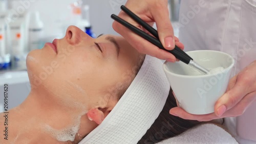 Woman receives facial treatment in salon. Therapist applies mask using brush. Atmosphere is focused on skincare and relaxation. Steps show careful application.