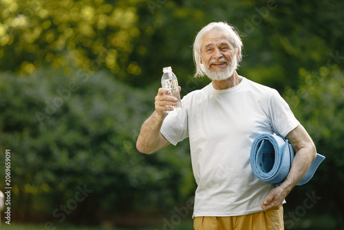 Senior man with exercise mat standing at park and holding bottle of water