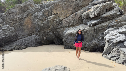 Fotografi Smiling young Hispanic woman walking barefoot on a sandy beach with dramatic coa