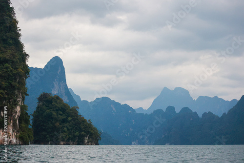 Moody tropical landscape of limestone karst mountains, forested cliffs, and a calm lake under cloudy sky in Thailand