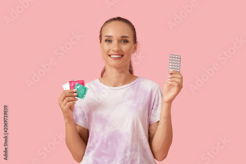 Young woman with condoms and contraceptive pills on pink background