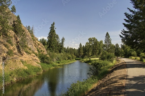 Sunny summer day view of the Trail of the Coeur d’Alenes passing beside a rocky cliff and small pond in the forest near Harrison, Idaho.