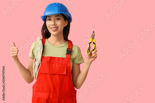Young female electrician with pliers and cables showing thumb-up on pink back...