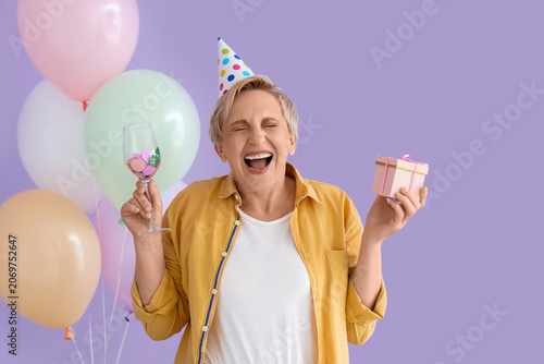 Mature woman with glass of confetti and birthday gift on lilac background