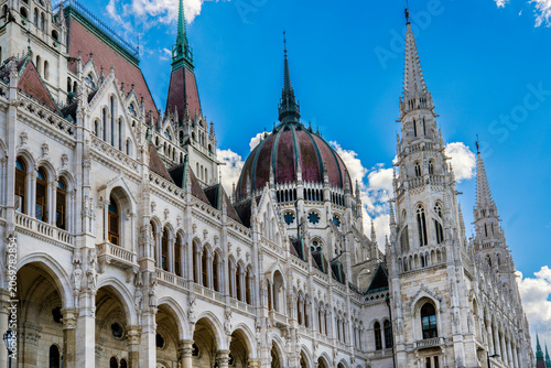 Detailed exterior view of the historic Hungarian Parliament Building in Budapest, Hungary, featuring its iconic dome, ornate facade and Neo-Gothic spires beneath a bright blue sky.