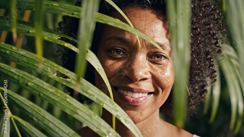Smiling middle aged black woman with hazel eyes looking through green palm leaves in tropical sunlight
