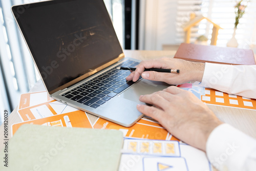 Man works at desk with laptop and blueprints. Hands tap keyboard while reviewing plans. Soft light glows behind workspace. Focused on task, calm and professional. Ideal for business, architecture

