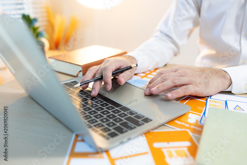Man works at desk with laptop and blueprints. Hands tap keyboard while reviewing plans. Soft light glows behind workspace. Focused on task, calm and professional. Ideal for business, architecture
