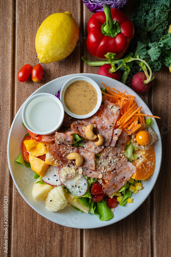 Fresh mixed vegetable and fruit salad bowl with crispy bacon, sesame dressing, and colorful ingredients on wooden background.