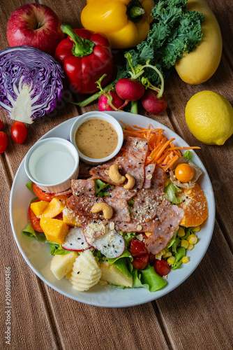 Fresh mixed vegetable and fruit salad bowl with crispy bacon, sesame dressing, and colorful ingredients on wooden background.