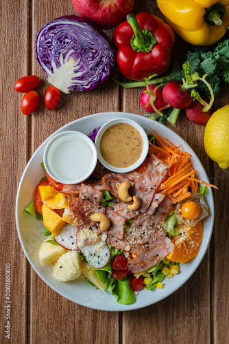 Fresh mixed vegetable and fruit salad bowl with crispy bacon, sesame dressing, and colorful ingredients on wooden background.