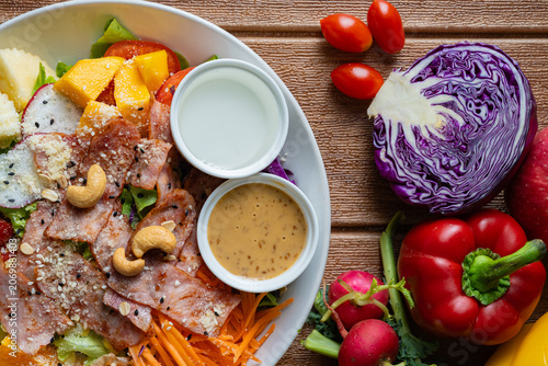 Fresh mixed vegetable and fruit salad bowl with crispy bacon, sesame dressing, and colorful ingredients on wooden background.