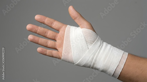 Injured hand wrapped in white medical bandage, wrist injury with gauze dressing, first aid treatment for sprain or fracture on gray background