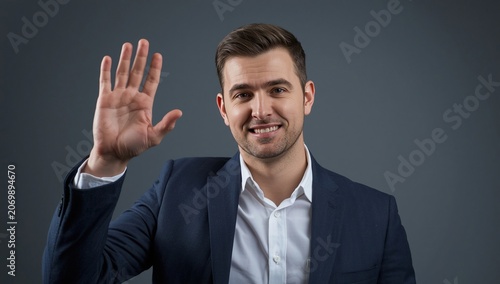 Smiling man in a navy suit waving his hand against a neutral dark gray background in a professional setting