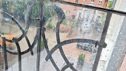 Rain covered window with decorative metal balcony railing overlooking an urban residential courtyard. Water droplets, blurred city details, and rainy weather create a moody atmosphere