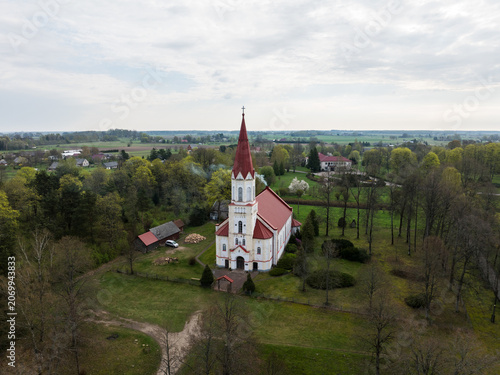 Aerial view of Rucava evangelical lutheran church with its red spire, surrounded by lush green trees and rural landscape in Rucavas pagasts, Dienvidkurzemes novad, Latvia.
