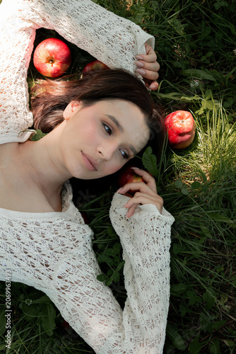 Creative portrait of woman in white lace outfit posing with red apples, balancing fruit on her head. Close up of girl with scraped, wounded knees covered with a green leaf in nature. Fairytale concept