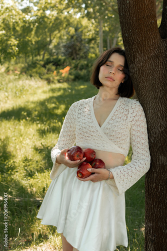 Creative portrait of woman in white lace outfit posing with red apples, balancing fruit on her head. Close up of girl with scraped, wounded knees covered with a green leaf in nature. Fairytale concept