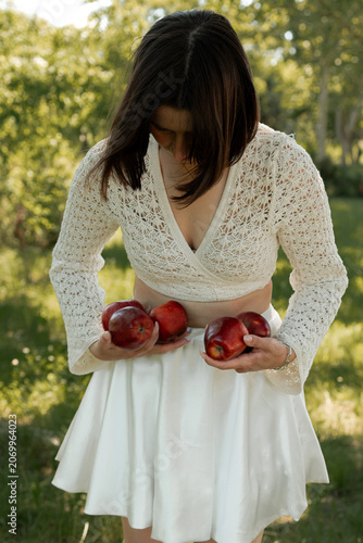 Creative portrait of woman in white lace outfit posing with red apples, balancing fruit on her head. Close up of girl with scraped, wounded knees covered with a green leaf in nature. Fairytale concept