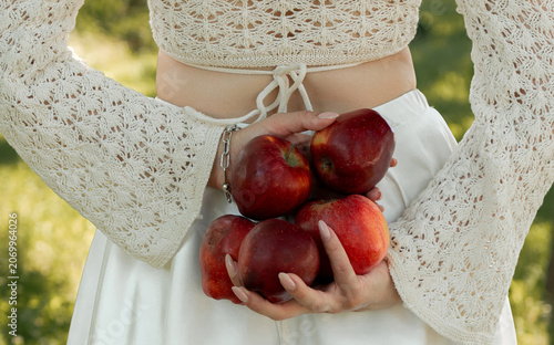 Creative portrait of woman in white lace outfit posing with red apples, balancing fruit on her head. Close up of girl with scraped, wounded knees covered with a green leaf in nature. Fairytale concept